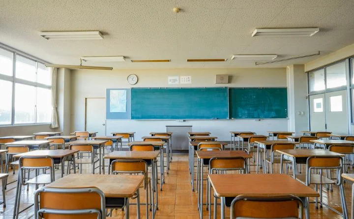 Pusta sala lekcyjna z rzędami drewnianych ławek i krzeseł, dużą zieloną tablicą i zegarem.