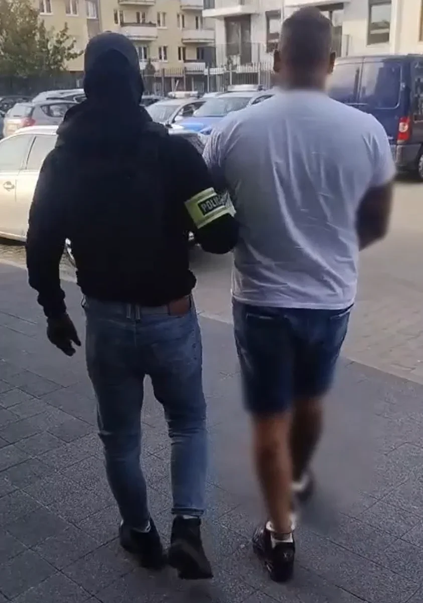 Funkcjonariusz w czarnej bluzie z opaską „POLICJA” prowadzi mężczyznę w białej koszulce ulicą miasta.