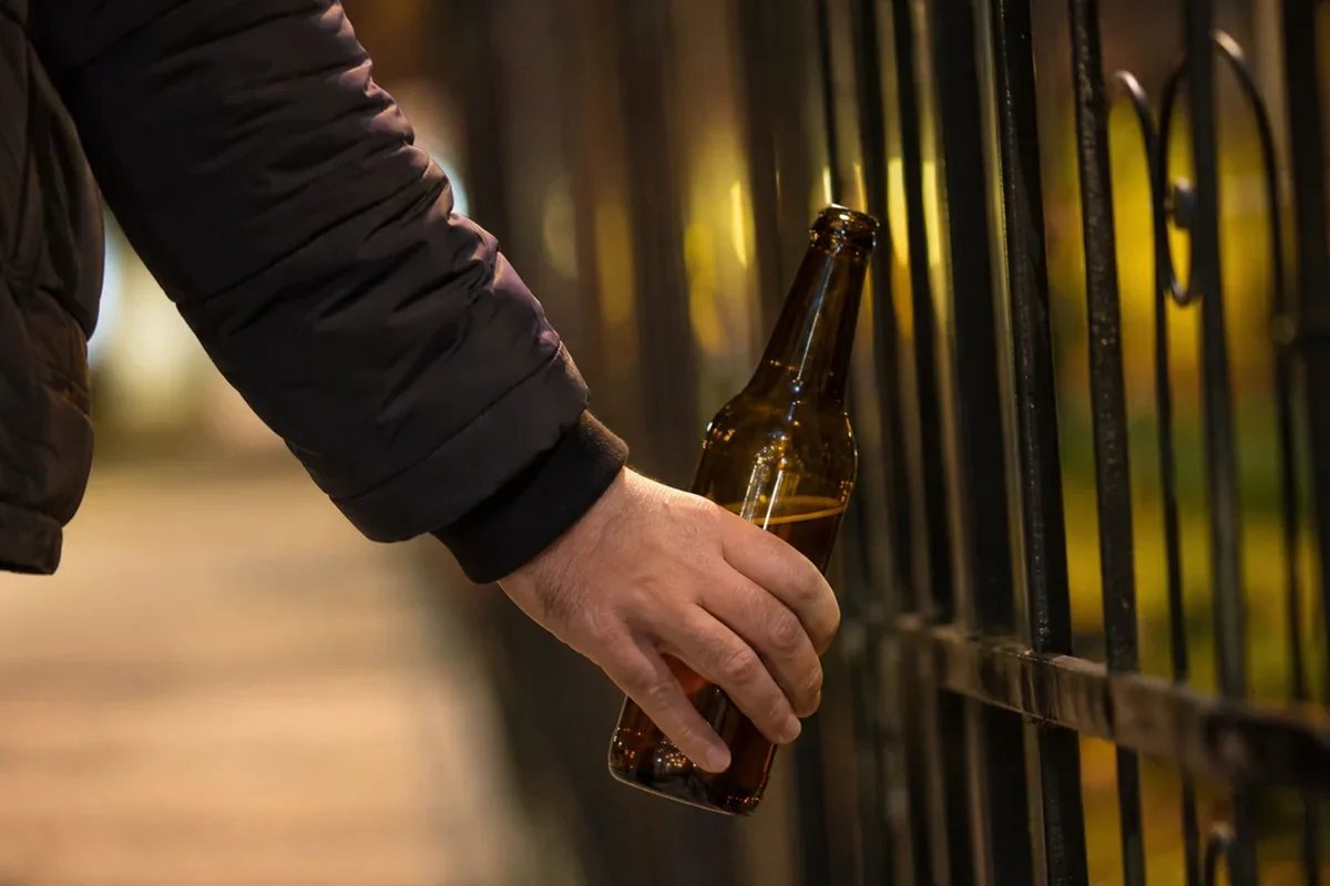 Mężczyzna w ciemnej kurtce trzyma w ręku butelkę piwa przy metalowym ogrodzeniu nocą.