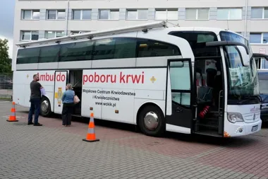 Mobilny ambulans do poboru krwi Wojskowego Centrum Krwiodawstwa i Krwiolecznictwa stoi na brukowanym placu przed budynkiem, przy którym czekają dwie osoby.