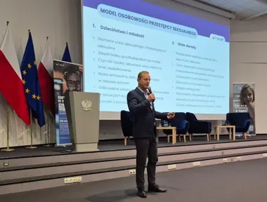 Mężczyzna w garniturze przemawia przez mikrofon na scenie konferencyjnej z dużym ekranem prezentacyjnym, flagami i pulpitami w tle.