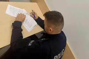Policjant w mundurze siedzi przy biurku i wypełnia dokumenty długopisem.