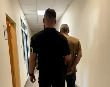 Funkcjonariusz służby mundurowej prowadzi zakuwanego w kajdanki mężczyznę korytarzem budynku urzędowego.