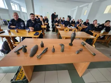 Policjanci w mundurach siedzą w sali podczas szkolenia, a na pierwszym planie na drewnianym stole leżą różne rodzaje niewybuchów i amunicji.