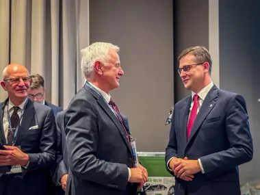 Dwaj mężczyźni w garniturach rozmawiają podczas formalnego spotkania konferencyjnego, w tle inni uczestnicy i baner z napisem „XXV-lecie”.