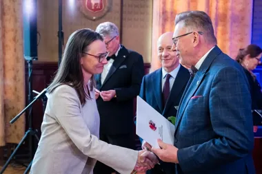 Kobieta w jasnym garniturze wręcza dokument mężczyźnie w niebieskim garniturze podczas oficjalnej uroczystości w reprezentacyjnym wnętrzu.