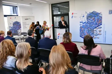 Mężczyzna przemawia z pulpitu podczas konferencji w sali z uczestnikami siedzącymi na krzesłach, obok stoją trzy kobiety, a w tle widoczne są rollupy promocyjne