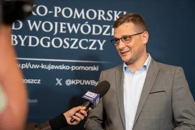 Mężczyzna w szarym garniturze udziela wywiadu dziennikarzowi z mikrofonem TVP3 Bydgoszcz przed ścianką Urzędu Wojewódzkiego w Bydgoszczy.