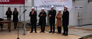 Grupa osób w kaskach budowlanych stoi na placu budowy podczas uroczystości, w tle baner generalnego wykonawcy.